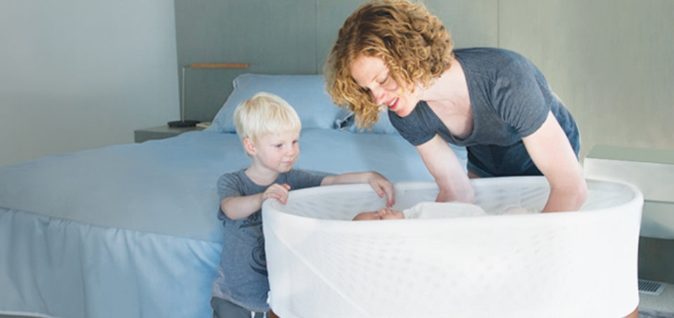 A woman checking on her baby inside a Snoo Smart Bassinet, with her older child watching beside her